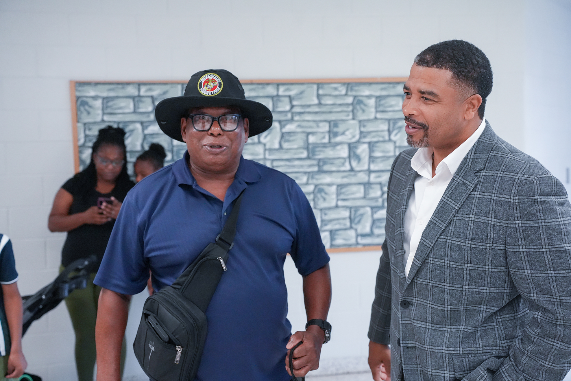 Two men stand conversing; one wears a hat and glasses, and the other wears a gray suit. A mural is on the wall behind them.