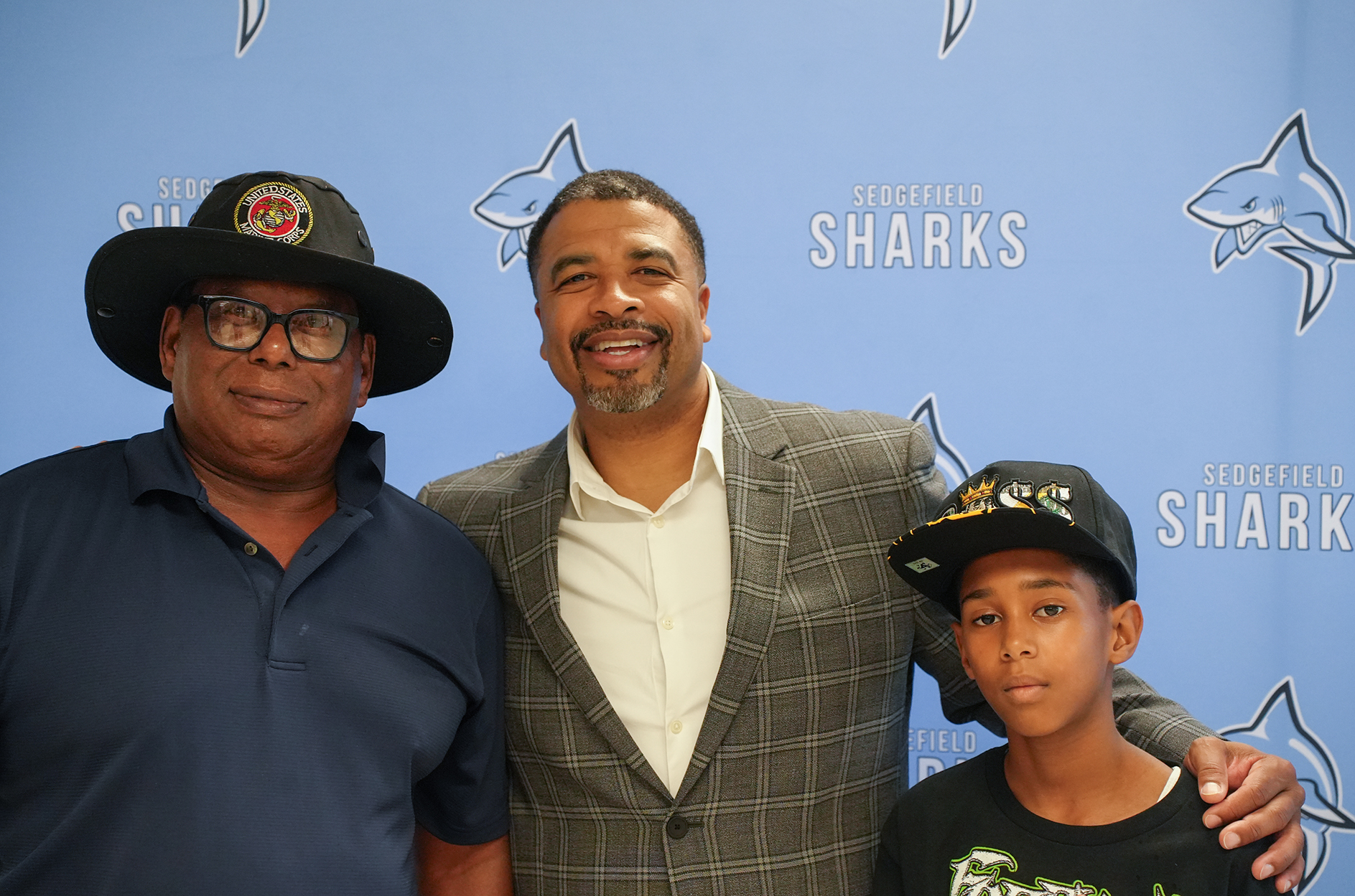 Three individuals in front of a wall with a logo. The man on the left wears a hat and glasses. The man in the middle wears a suit. The boy on the right wears a cap.