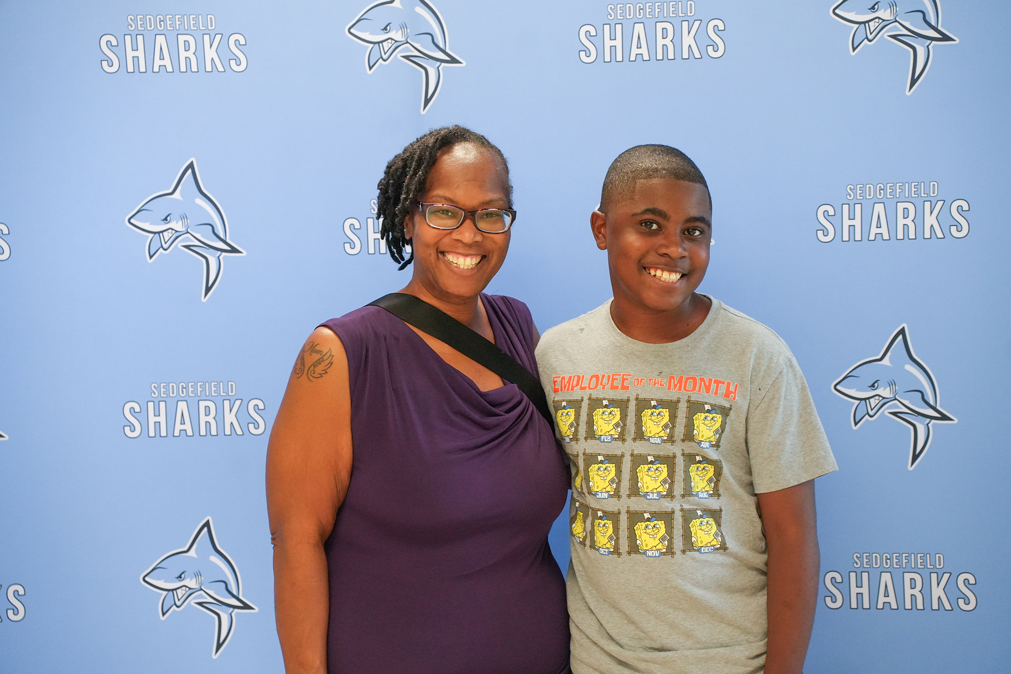 Two people stand close together against a blue wall with cartoon sharks, smiling for a photo.