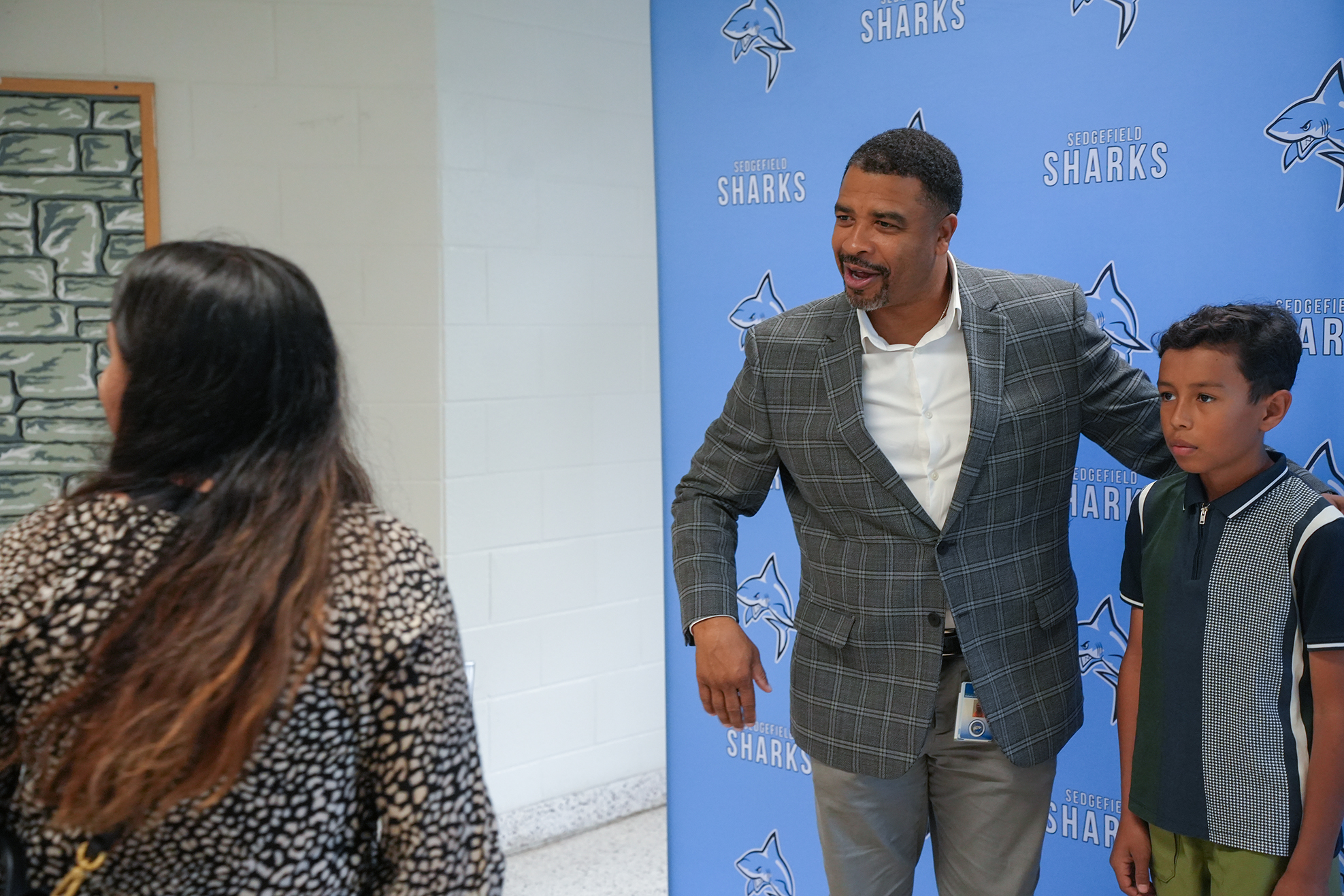 Man in formal suit stands with arm around boy, facing woman with long hair in hallway with blue Sharks backdrop.