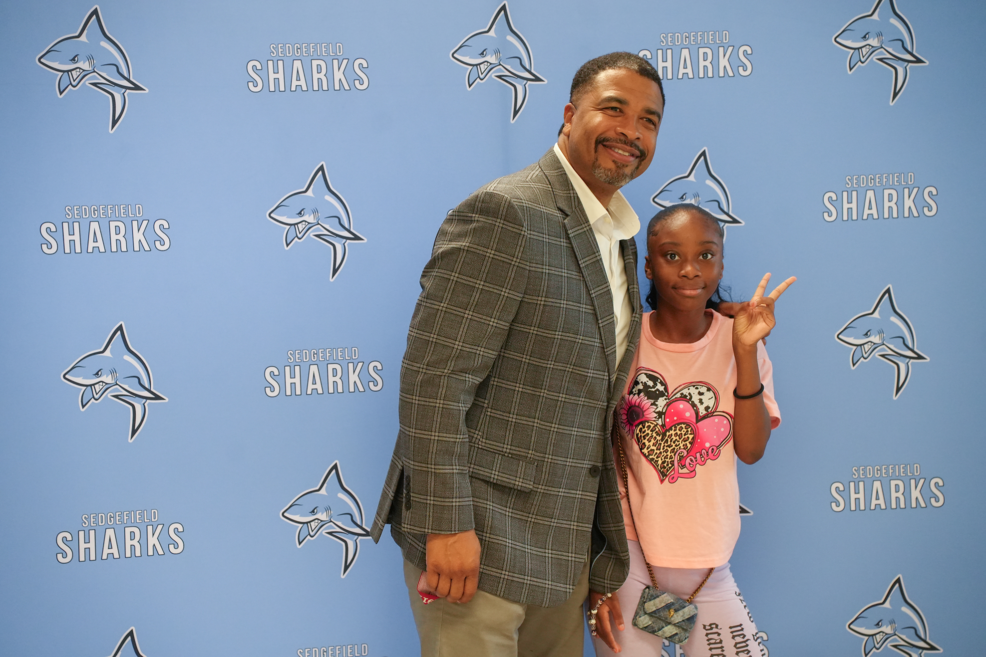 A man and a young girl pose together in front of a blue wall with "Sharks" logos.