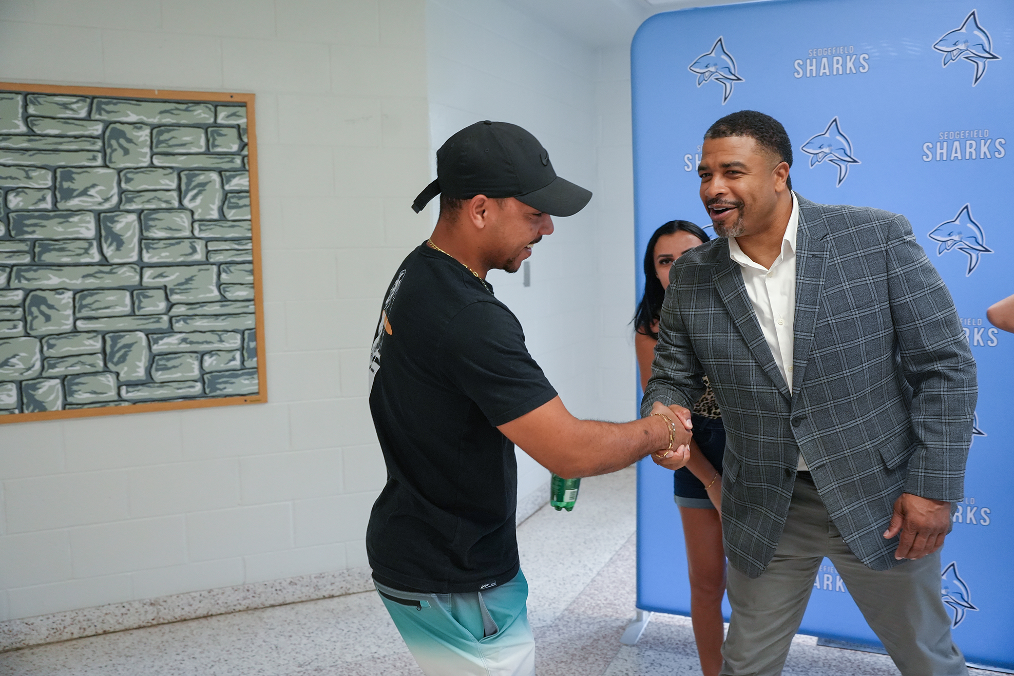 Two men shake hands in front of a mural. One wears a black hat and shirt, the other a gray suit. A woman stands behind the suited man.