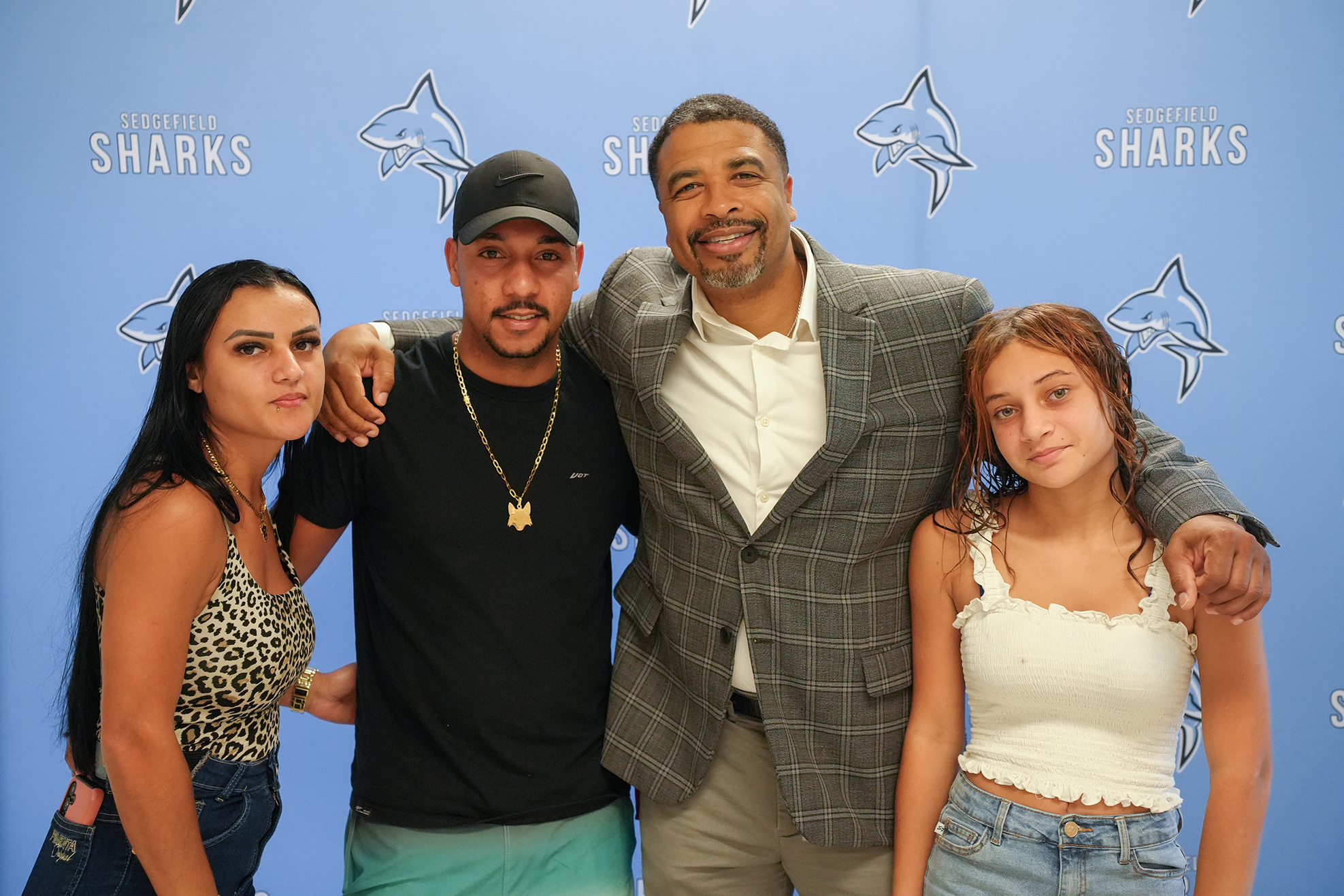 Four people pose together. Two adults and two girls. Behind them is a blue background with white sharks.