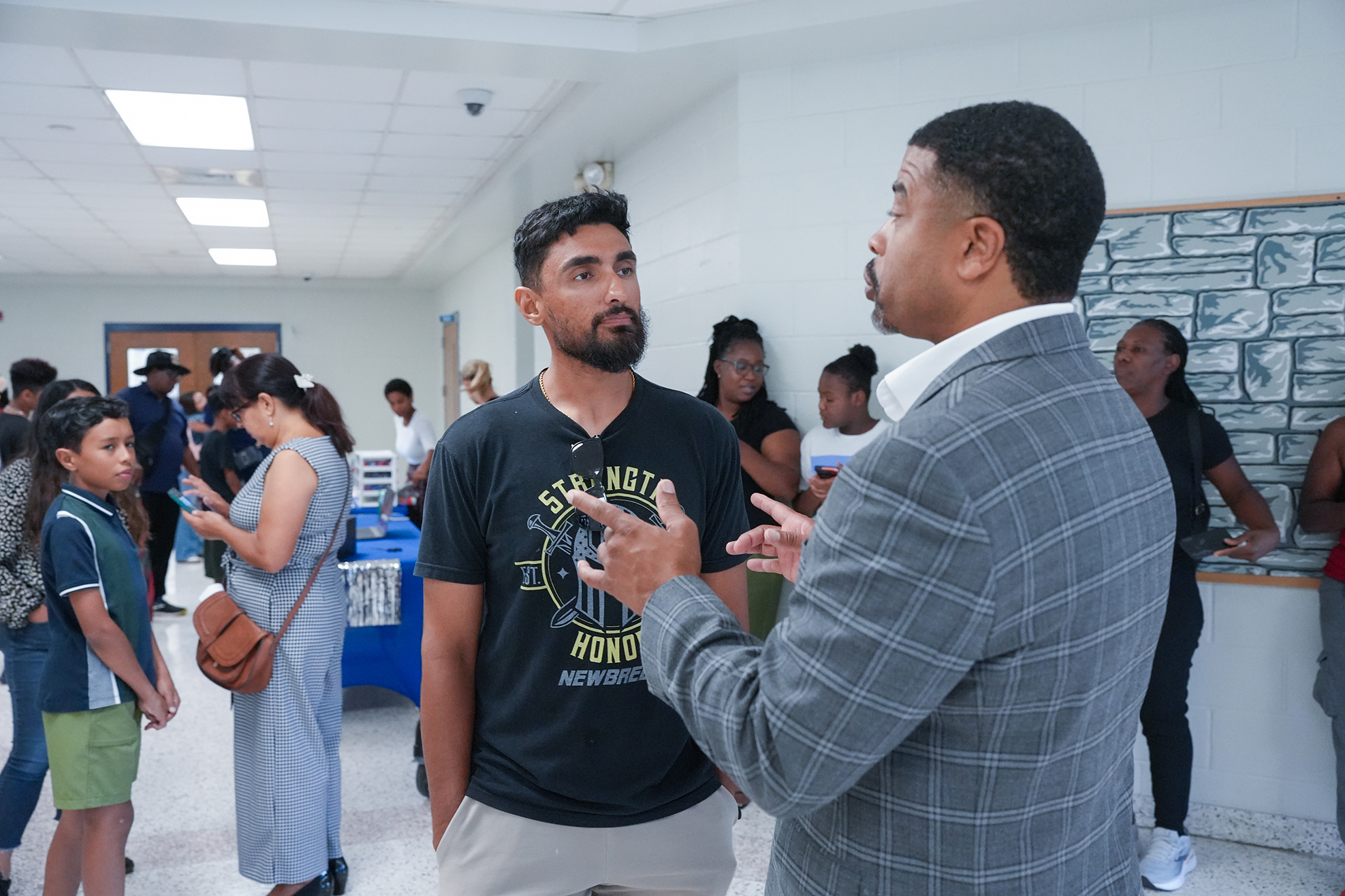 A man in a gray suit speaks to a man in a black t-shirt. They stand in a hall with tables.