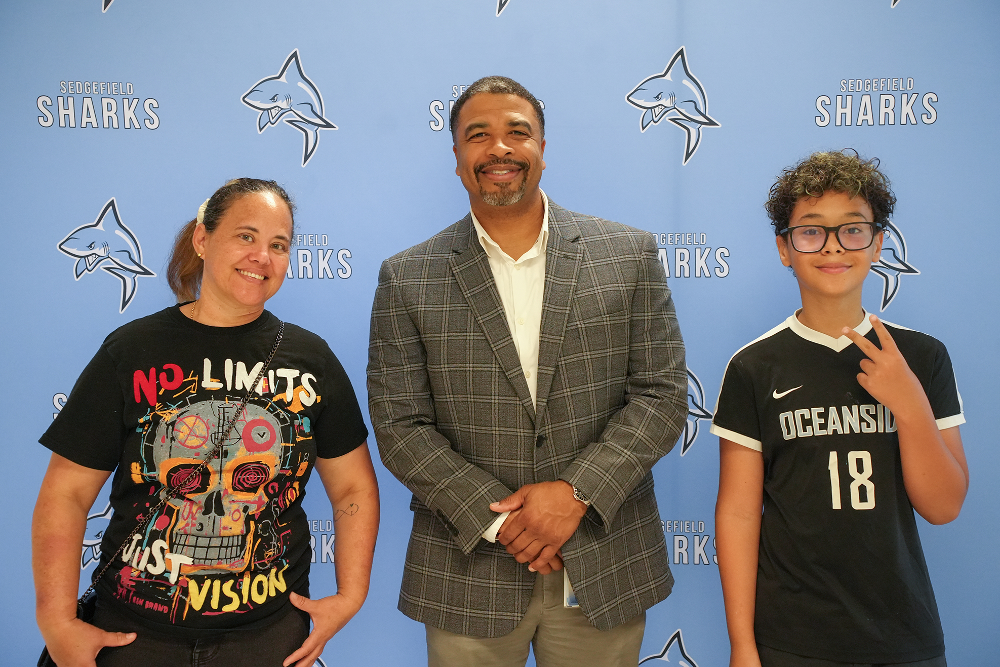 Three individuals stand together in front of a blue wall with a shark logo. One wears a skull-themed black shirt, another a gray blazer, and the third a black sports jersey.