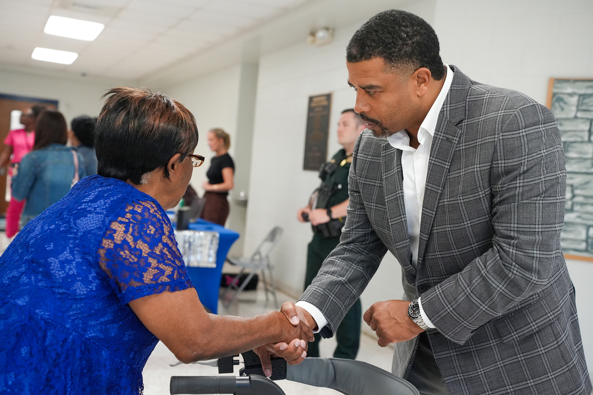 A man in a gray suit shakes hands with a woman in a blue dress. Behind them, people mill around, and a security officer stands near a wall.