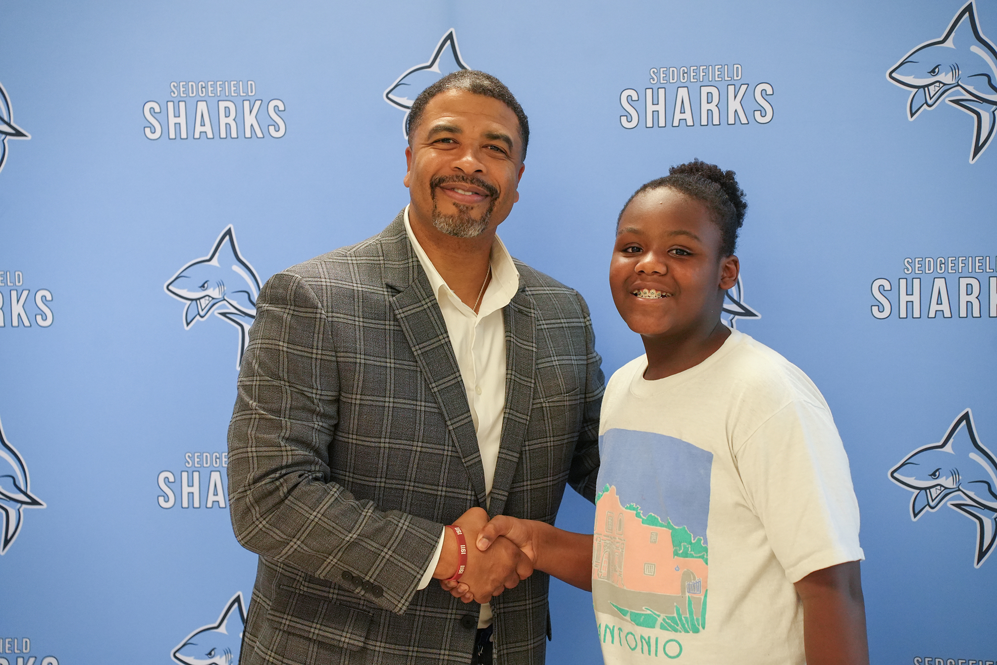 A man in a gray suit and a young girl in a white T-shirt shake hands in front of a blue wall.
