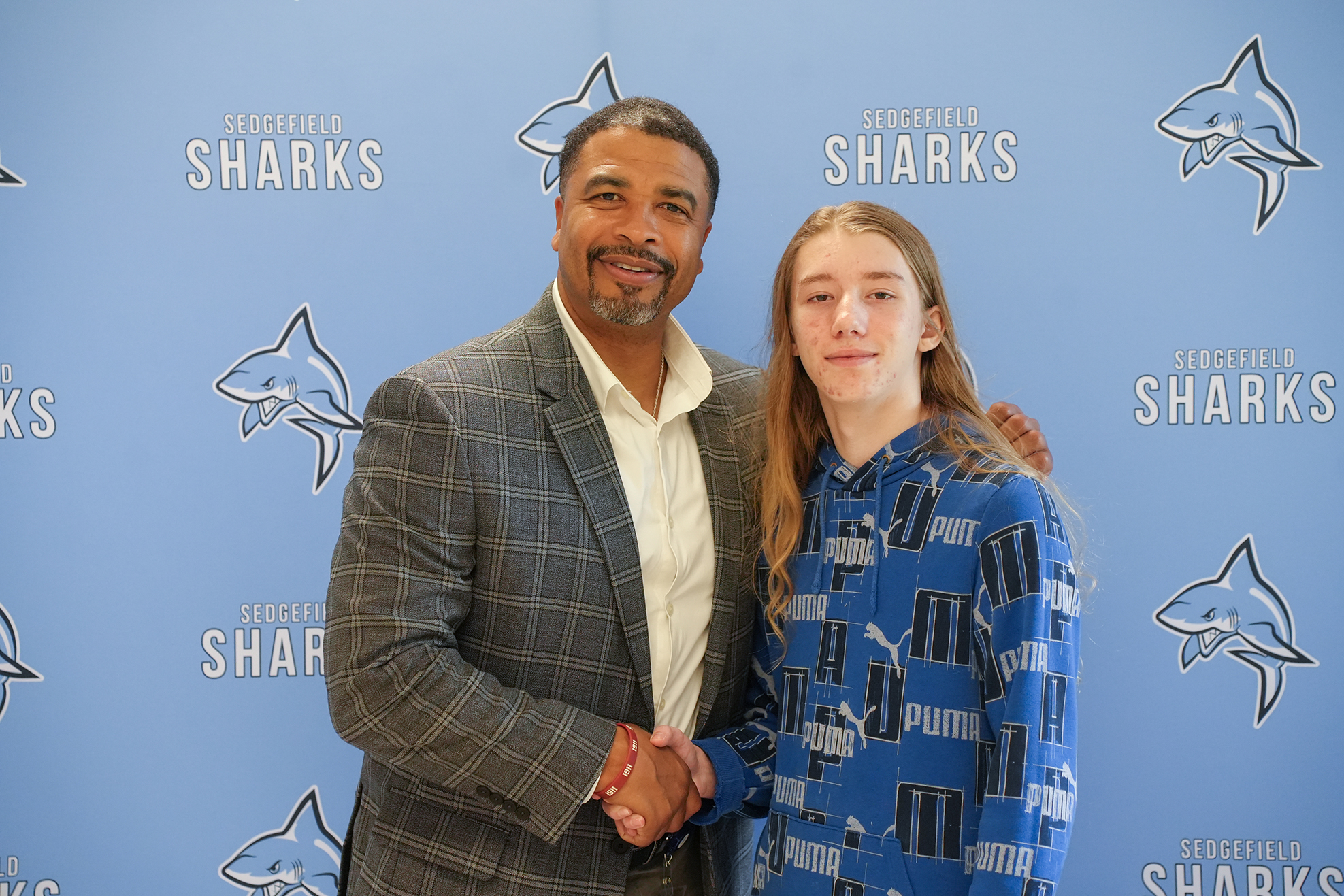 A man and a girl stand in front of a blue wall with shark graphics, shaking hands.