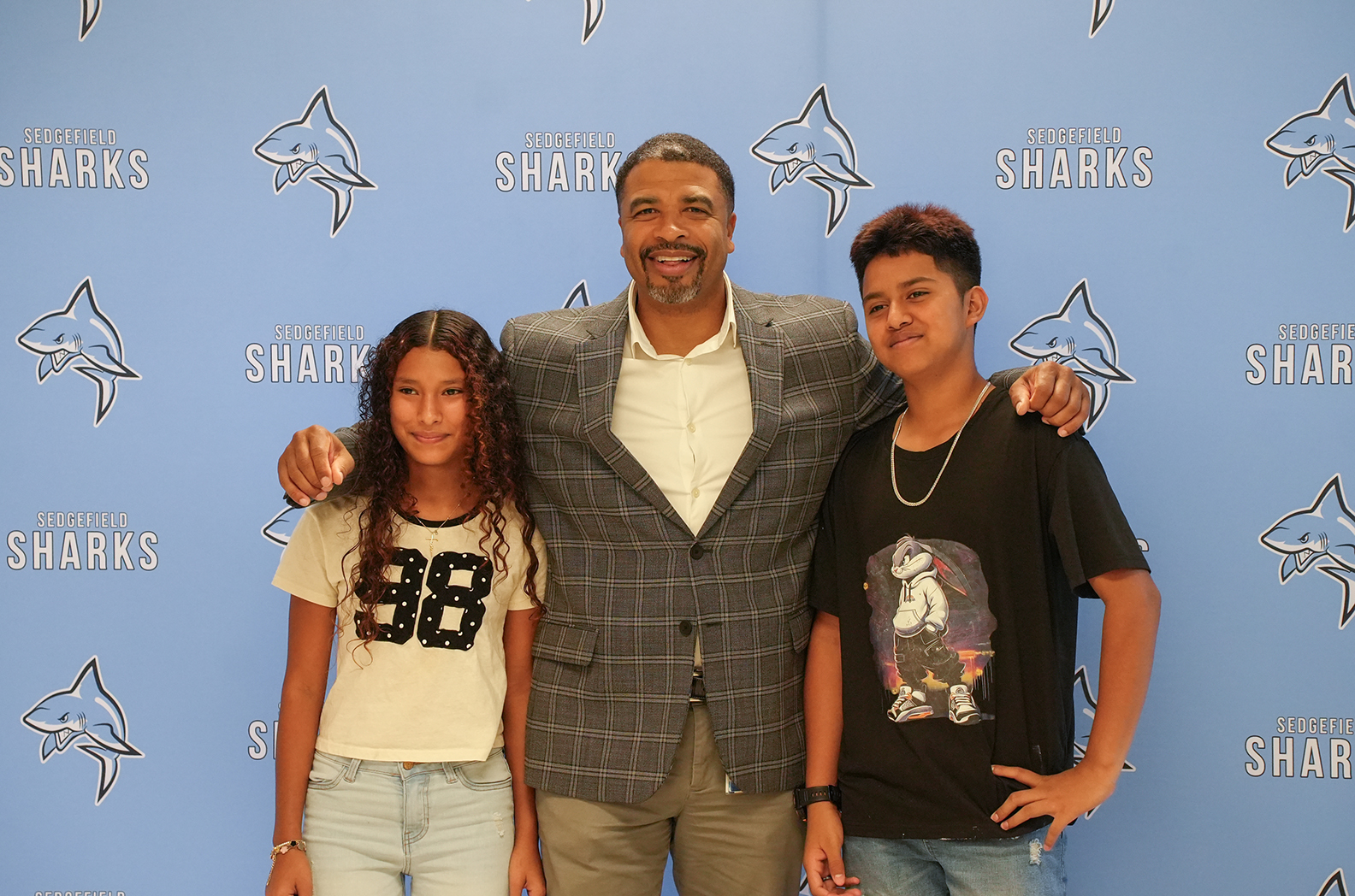 Three people pose for a photo against a blue backdrop with "Sharks" text. The man has his arms around two children.