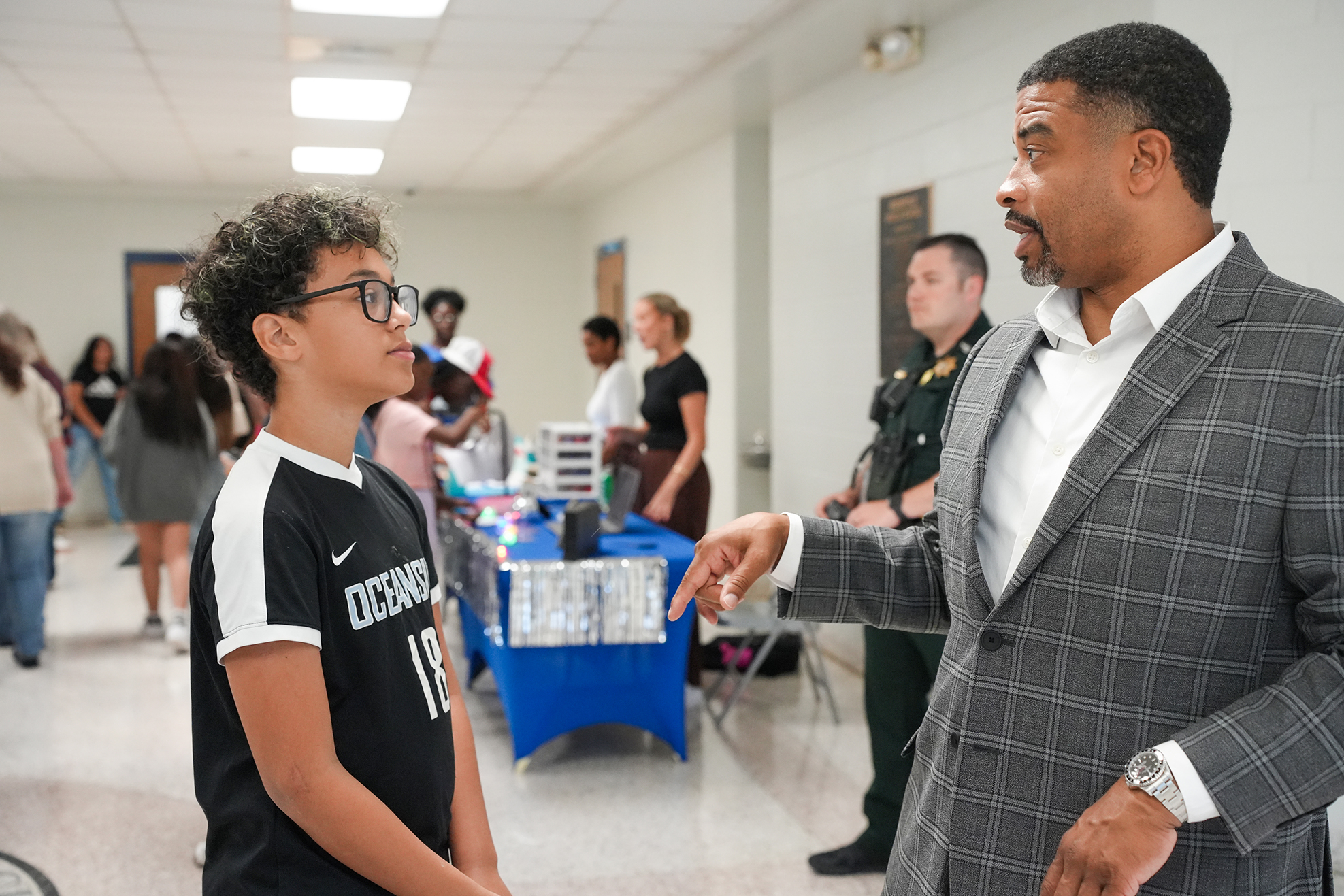 A man in a suit talks with a young boy wearing glasses. Behind them, people walk in a hallway with tables.