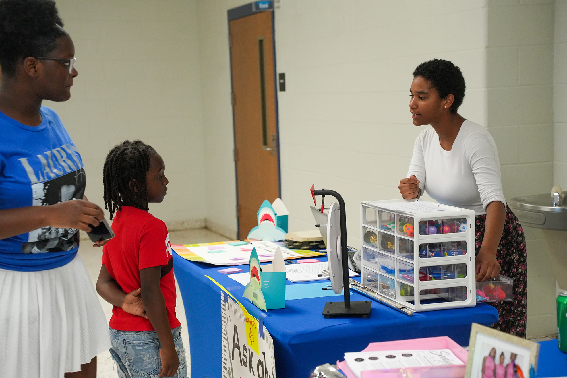 Three people in a classroom. A young girl speaks with an adult. Another girl looks at the adult.