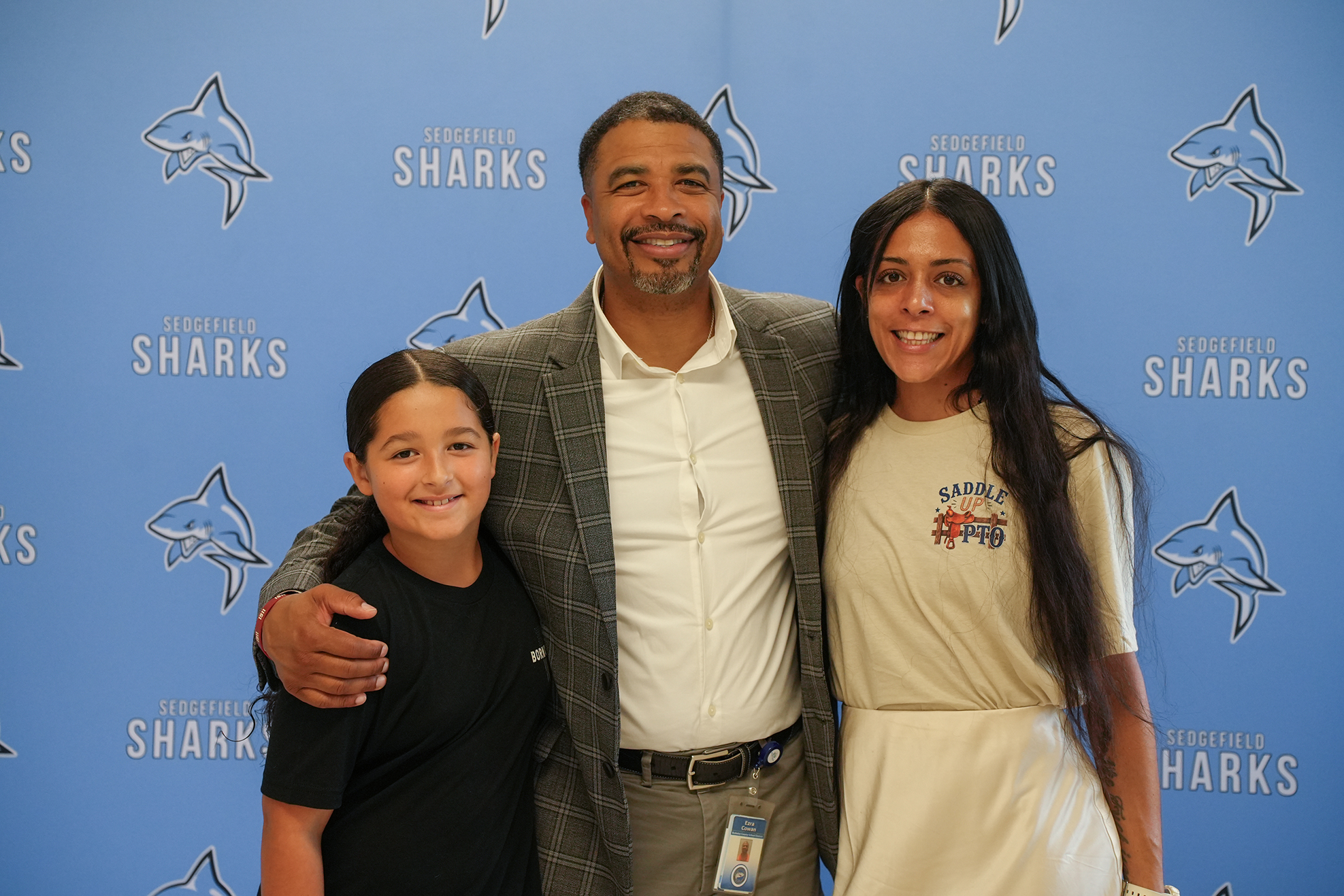 Three people stand together in front of a wall with blue and white shark logos. The man has his arm around two young girls.