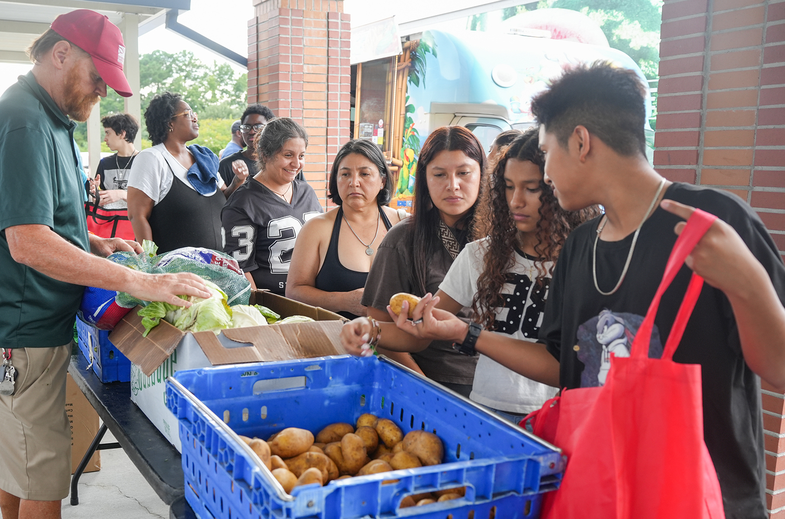 People gather around a table with produce in baskets. A man in a cap hands out food.