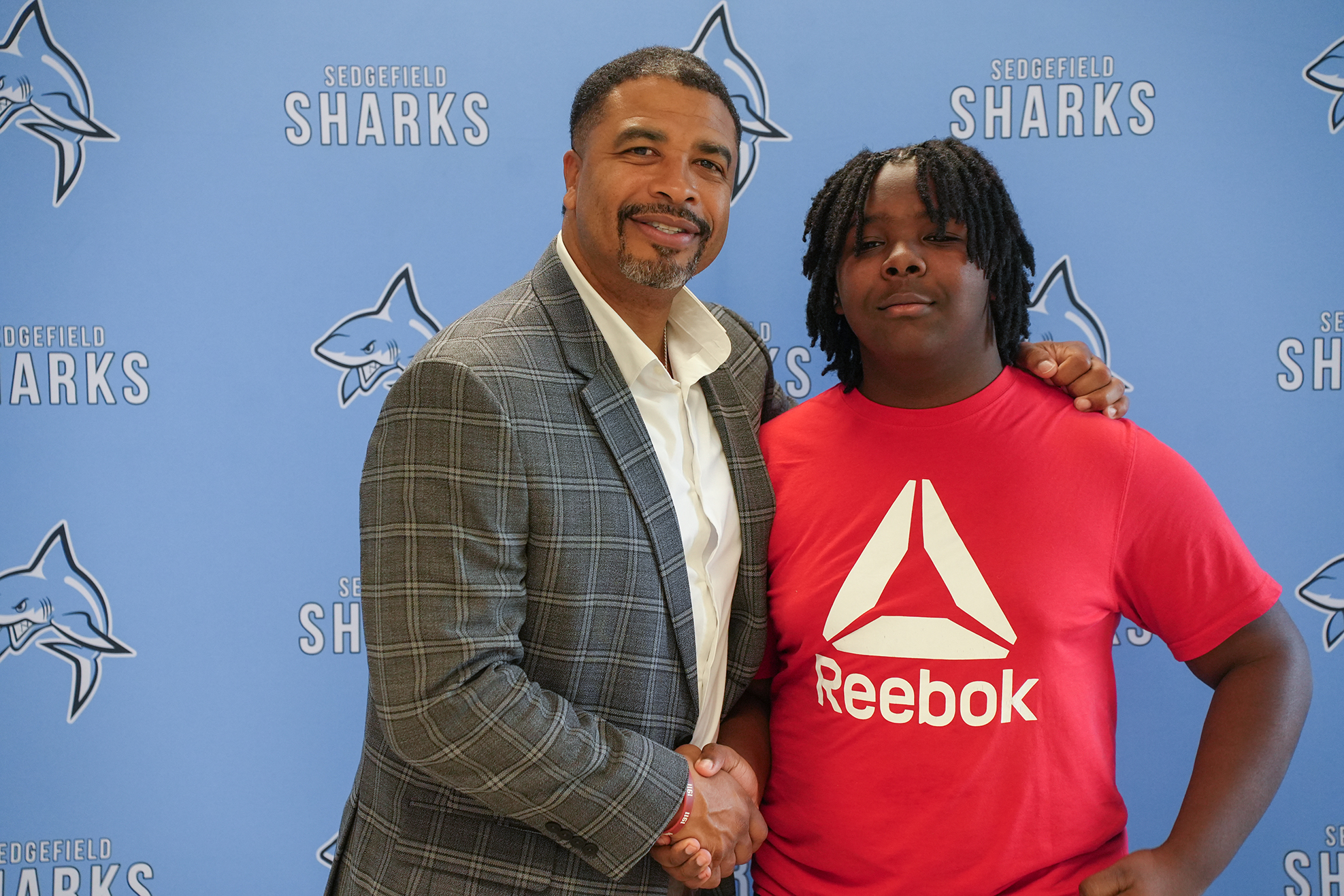 Two people, one in a Reebok shirt, standing and smiling in front of a blue wall with sharks logos.