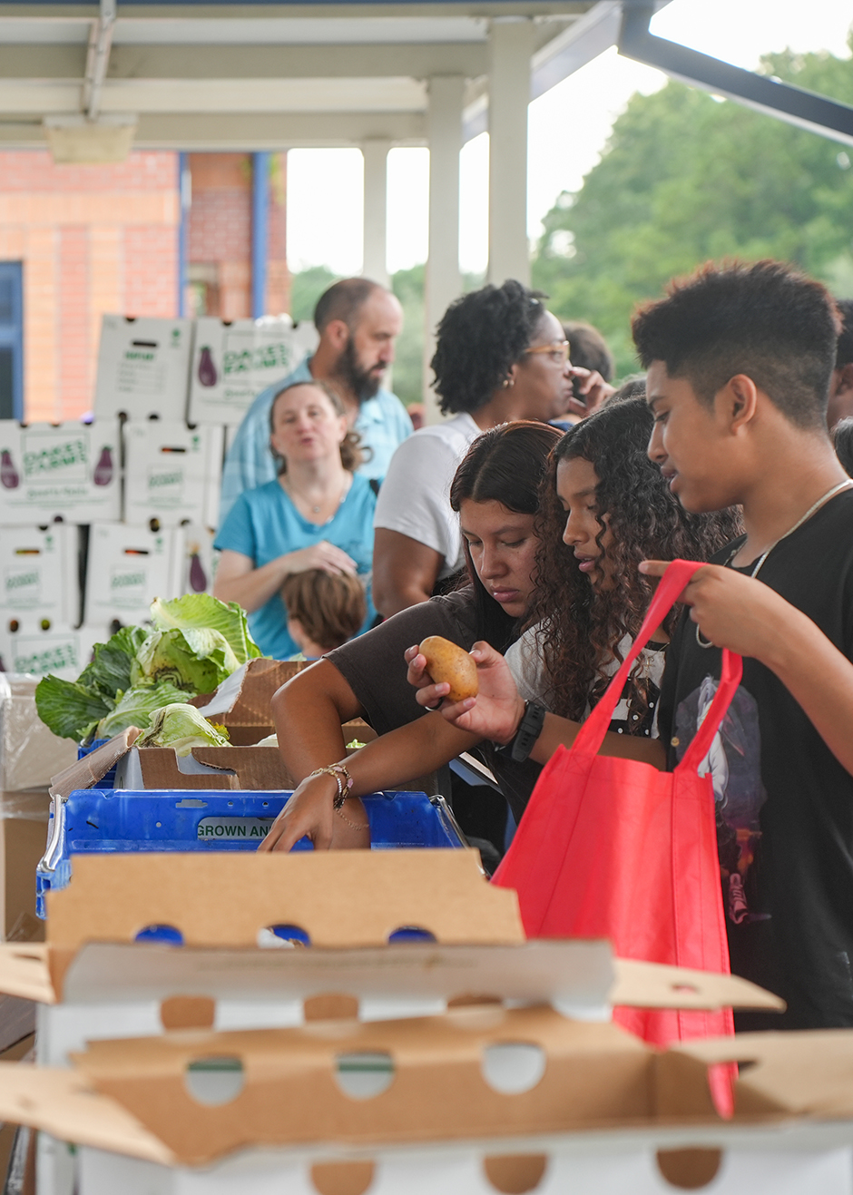 Group of people picking produce from boxes. Some people hold apples. A man holds a red bag.