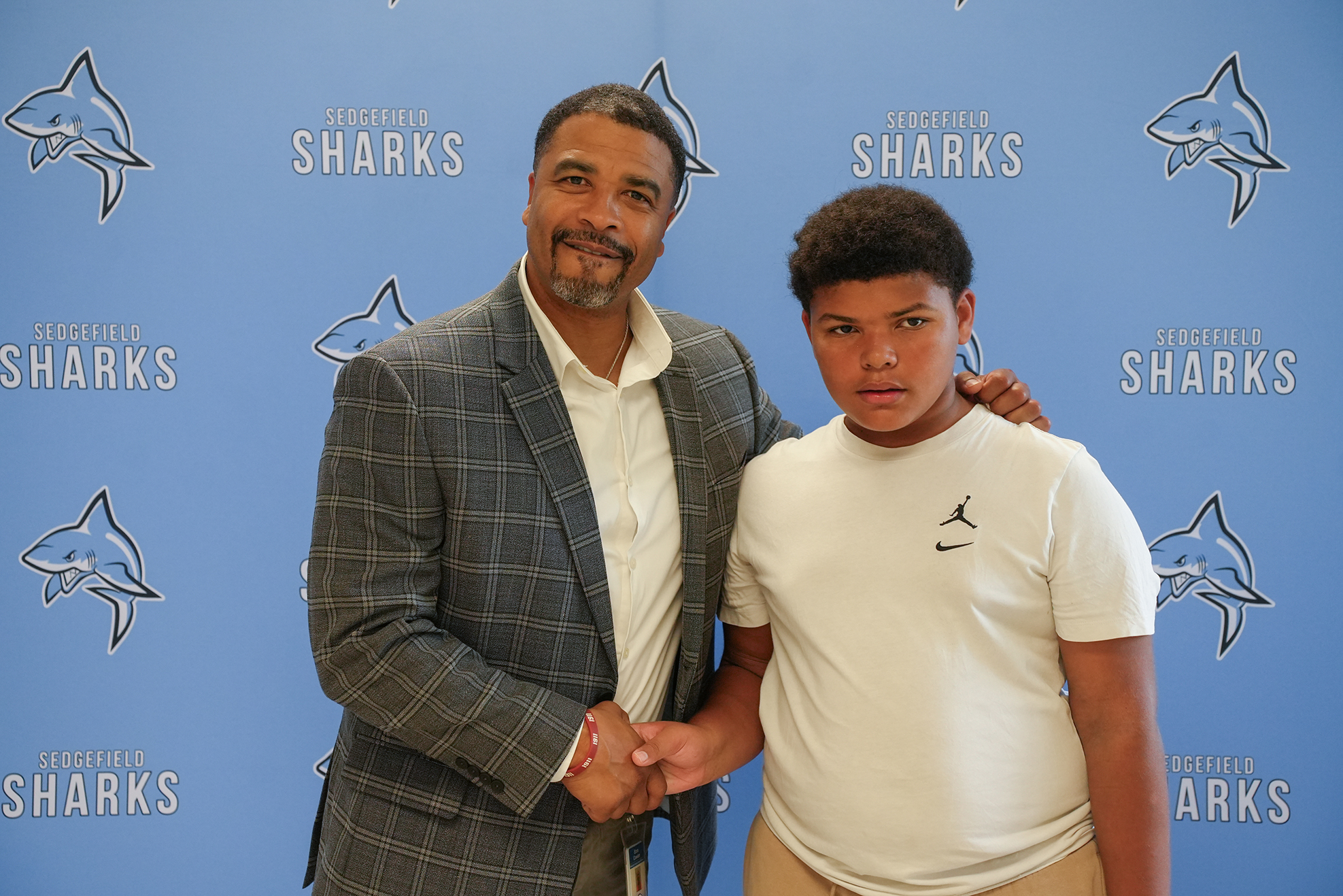 A man and a young boy standing together, smiling. The man wears a suit, the boy a white T-shirt.