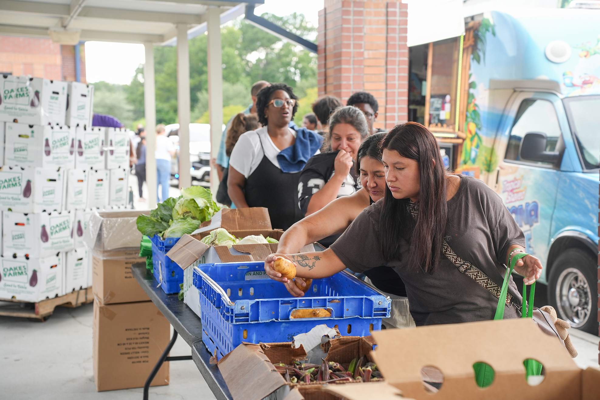 People at an outdoor market. A woman holds food in her hand. Boxes of produce on tables.