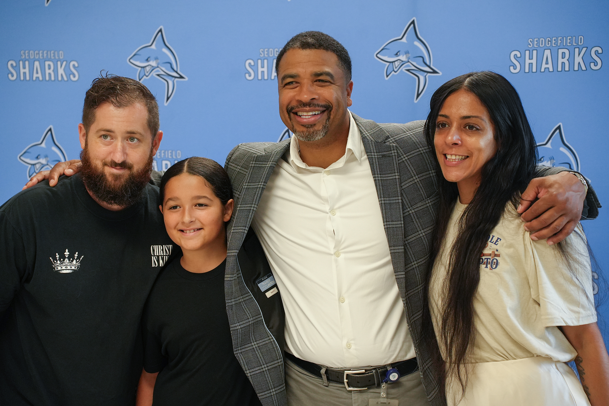 Four people pose for a photo. The two adults are smiling, and one holds a child. They stand before a wall with sharks and text.