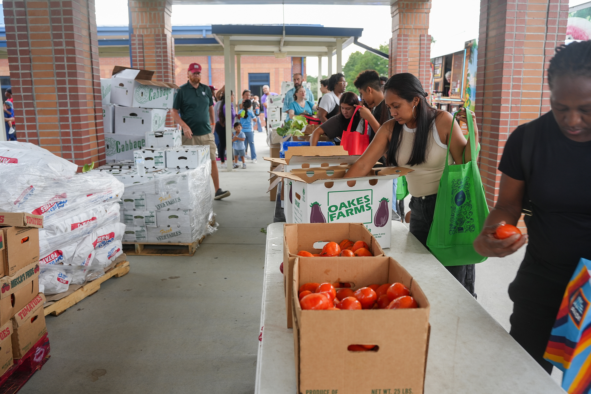 Covered market with multiple people shopping. Piles of produce in boxes. A woman holds a tomato.