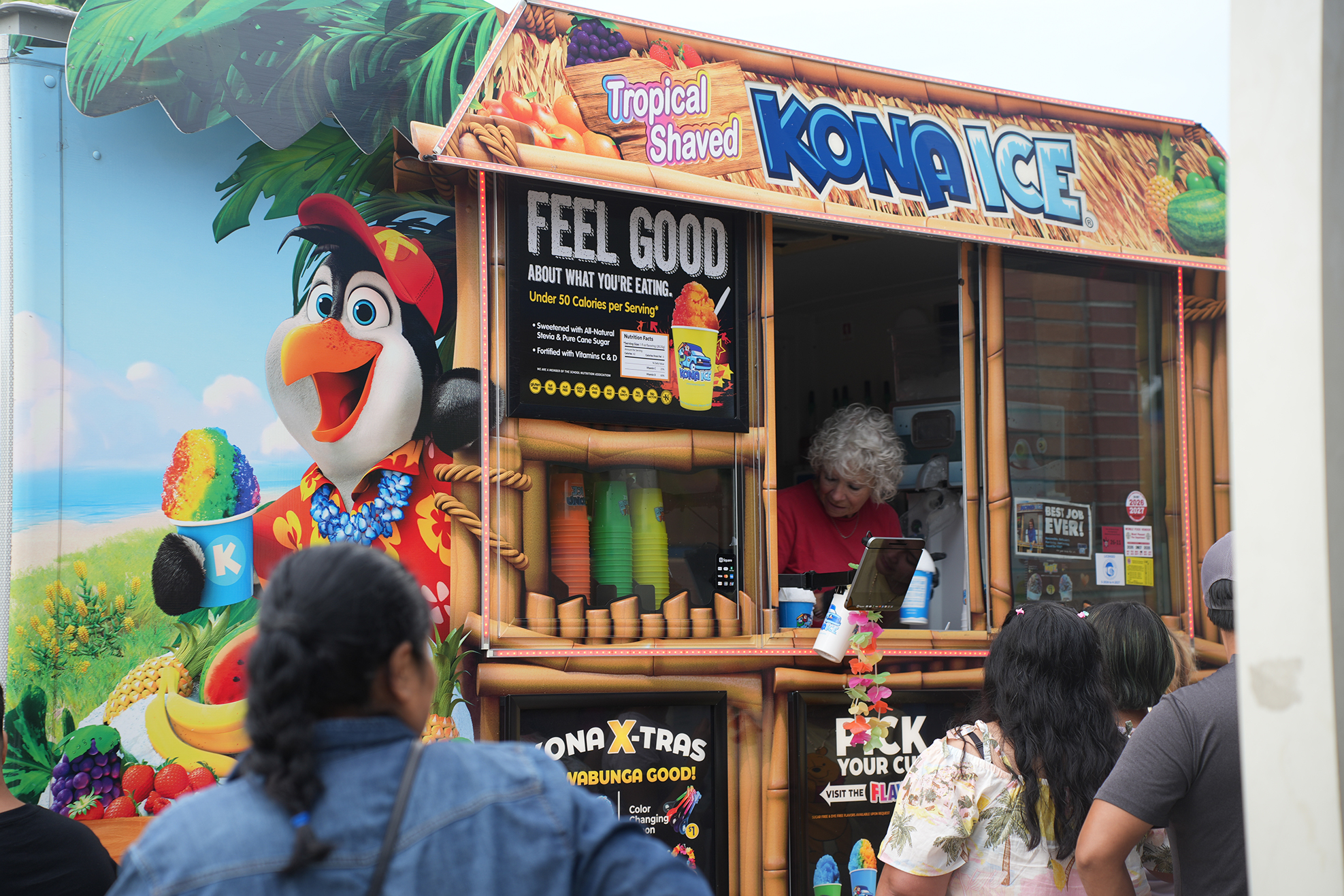 A Kona Ice truck is surrounded by people. A woman inside serves customers. The truck is painted with a tropical scene and a penguin.