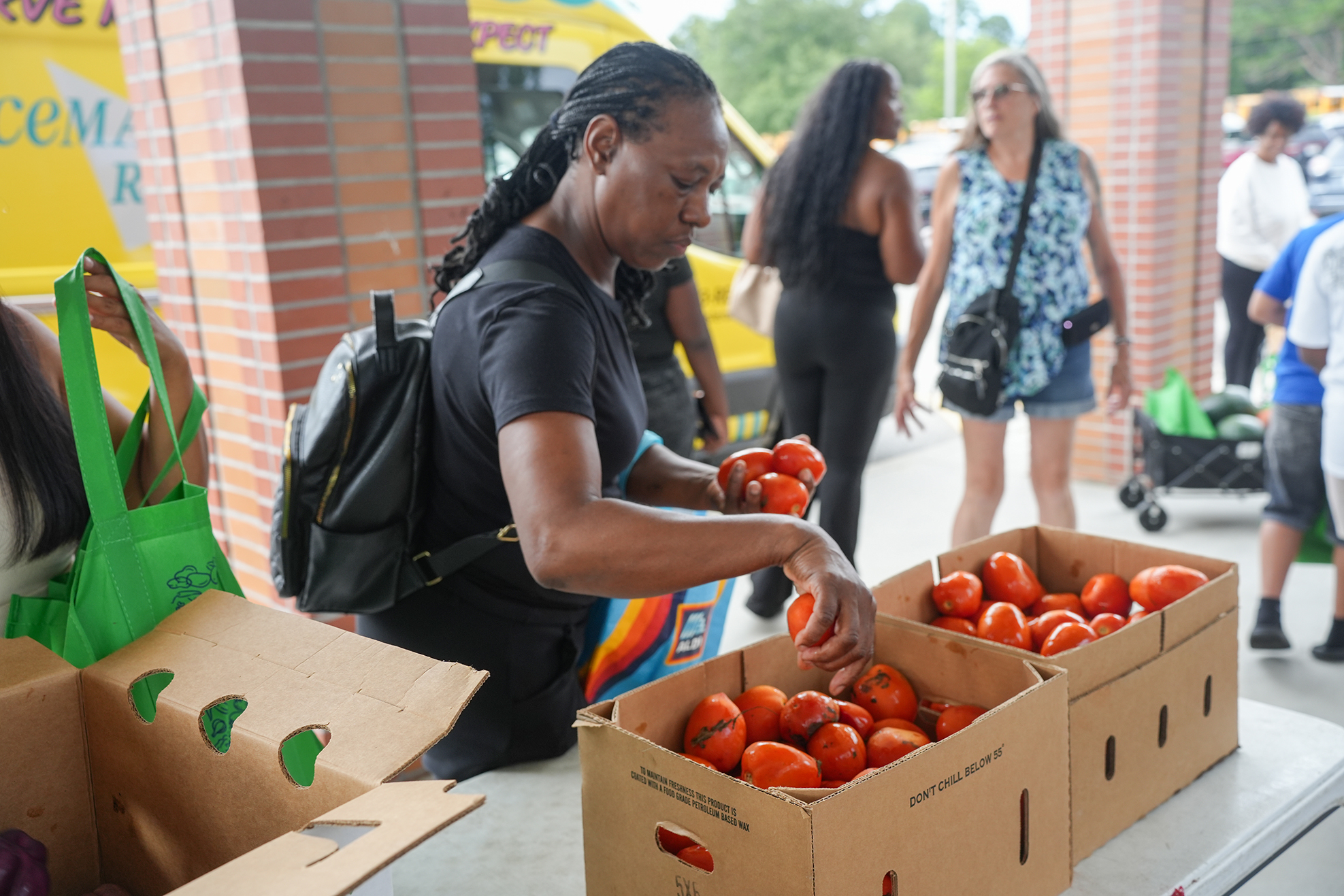 A woman selects tomatoes from a box while others walk by. One holds a green bag.