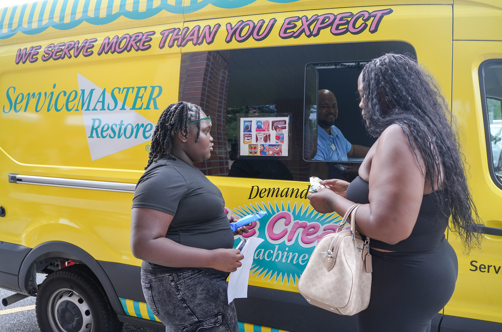 Two women stand outside a yellow food truck with "ServiceMaster Restore" and "Demand Cream" on it.