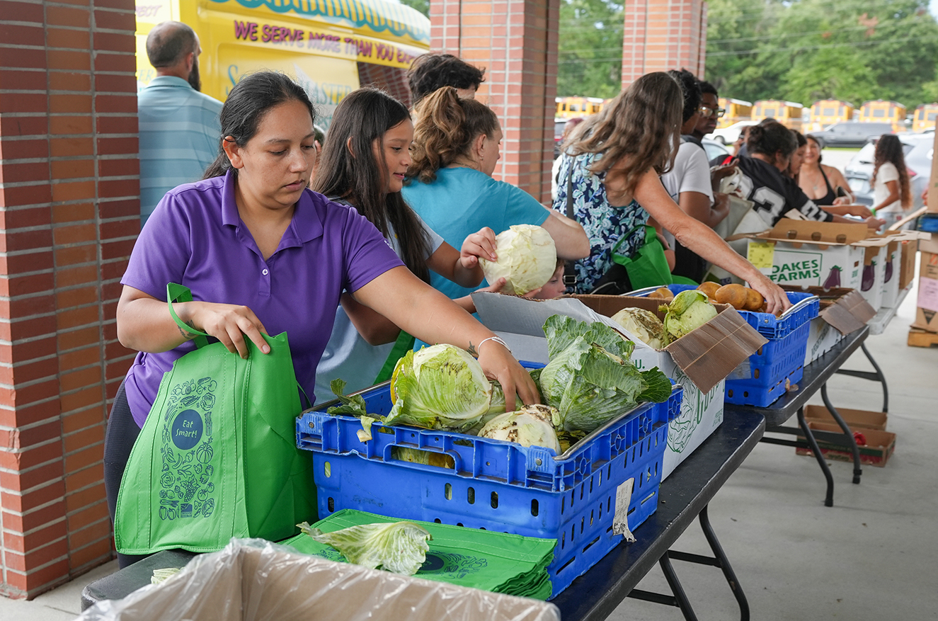 Women gather around a table filled with fresh vegetables, with some holding bags and others picking up produce.