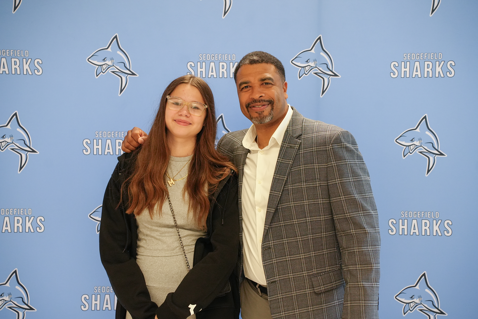 A woman with glasses and a man with a plaid jacket pose together in front of a blue backdrop with shark graphics.