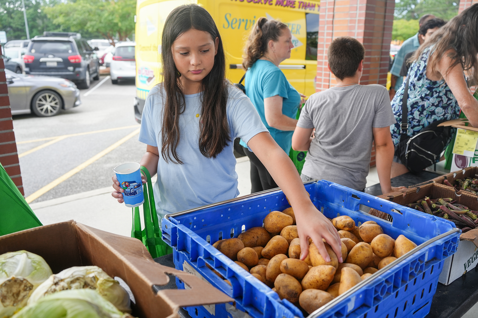 Children select produce at a community food distribution. A girl holds a potato. Vegetables are in front of them.