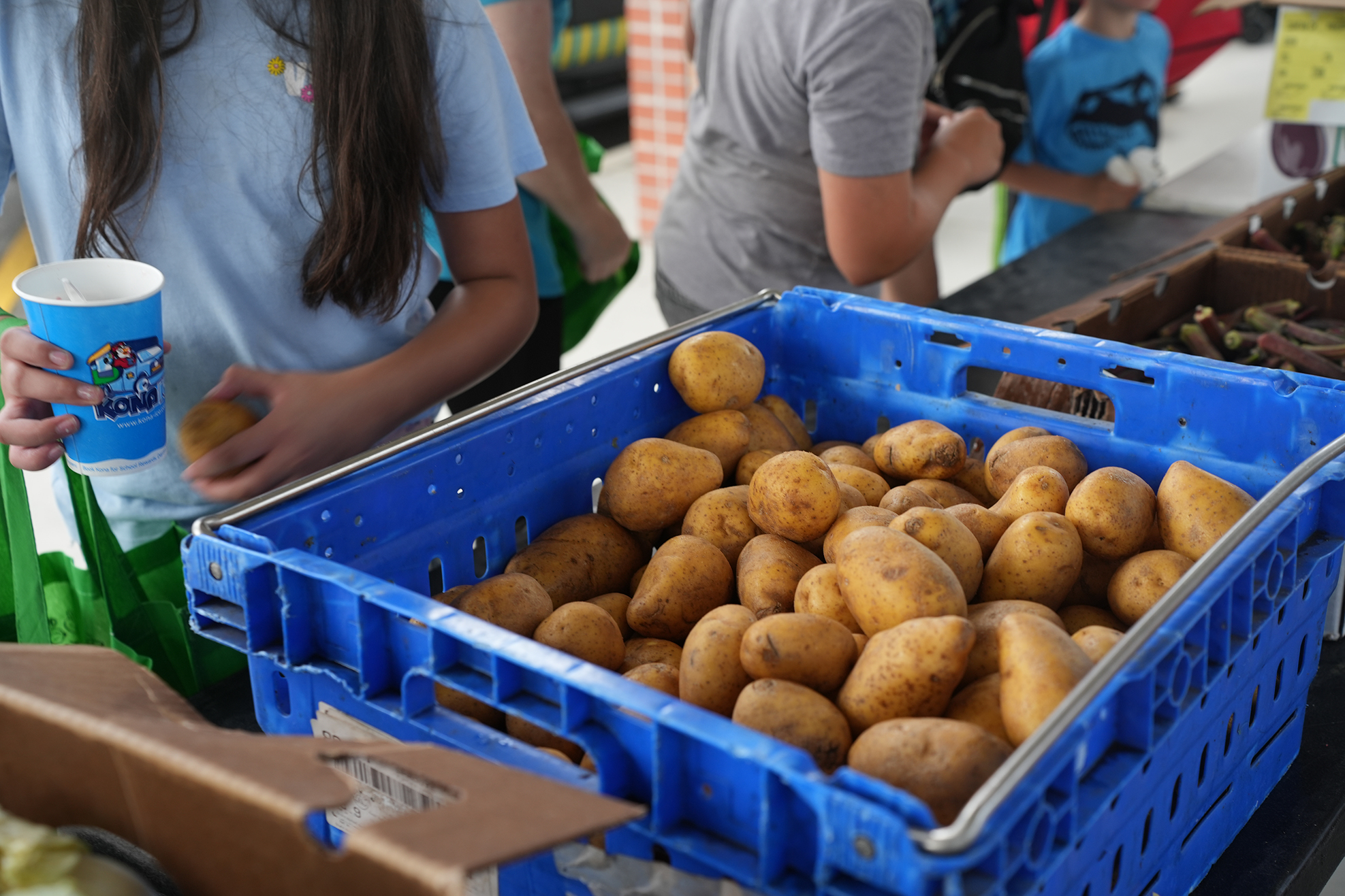 A person holds a cup in front of a blue basket of potatoes, with others shopping in the background.