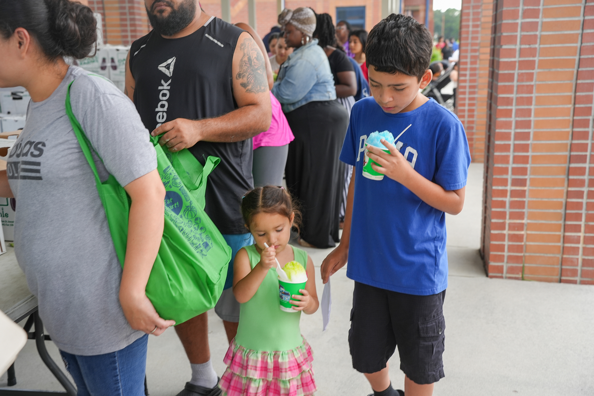 A group of people, including a young girl and a boy, stand in line at an event with brick walls in the background.
