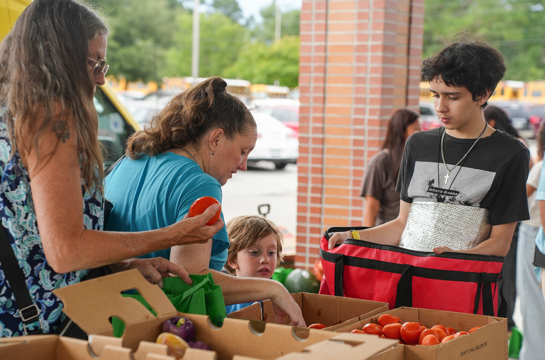 People standing around boxes of vegetables. One woman holds an orange. A boy holds a red bag.
