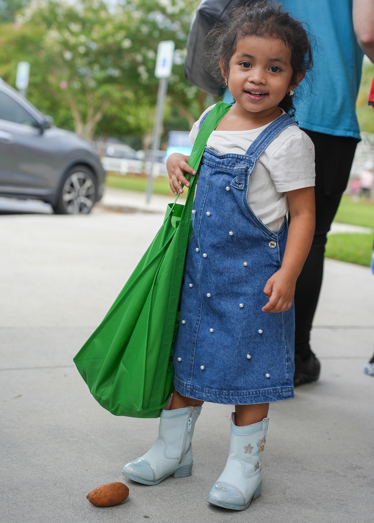 Young girl in a denim dress with pearl buttons, white shirt, and blue boots, holding a green bag, standing on a sidewalk.
