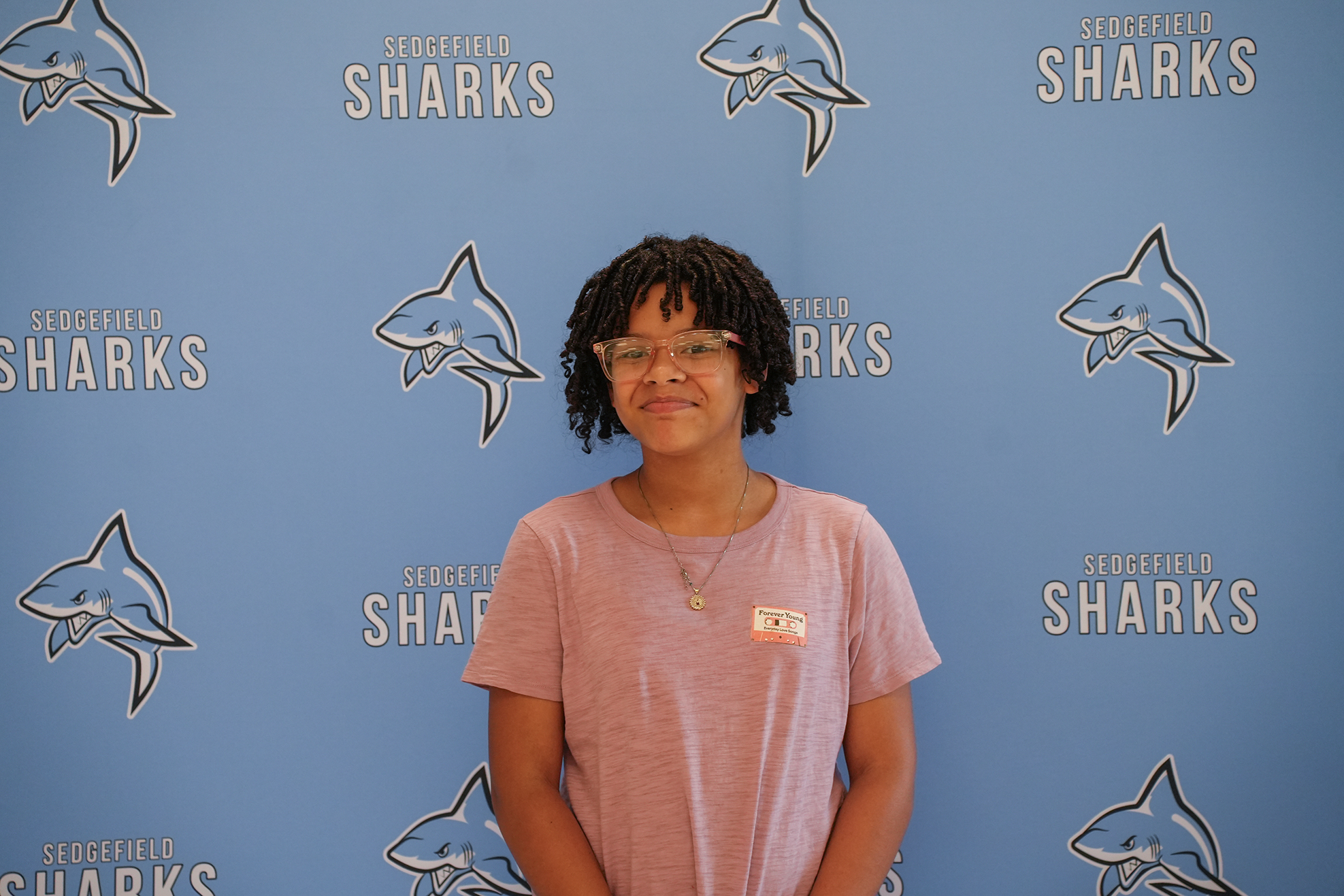 A young person with glasses and a pink shirt stands before a blue backdrop featuring several shark images.