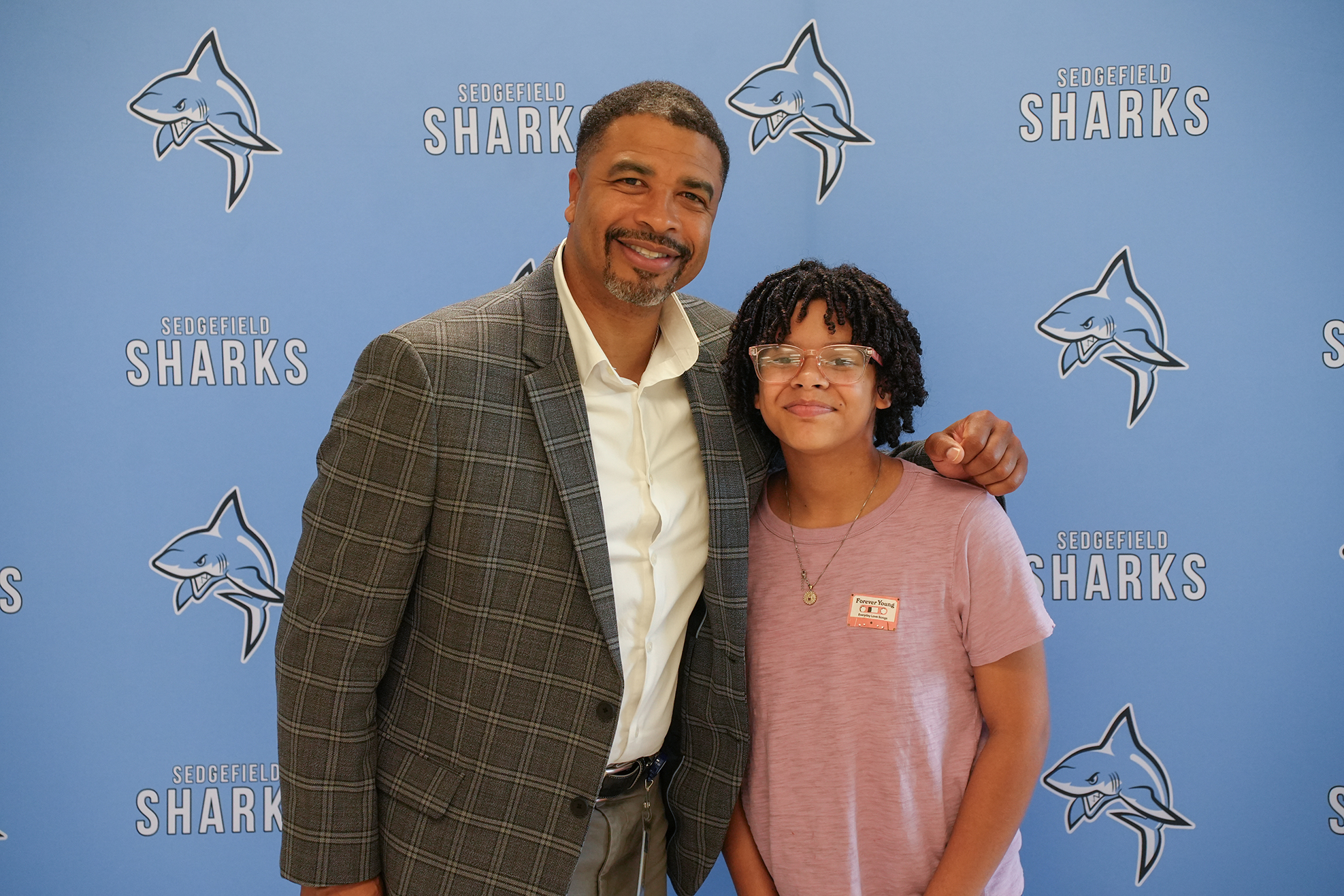 Man and child standing next to each other in front of a blue wall with sharks logo.