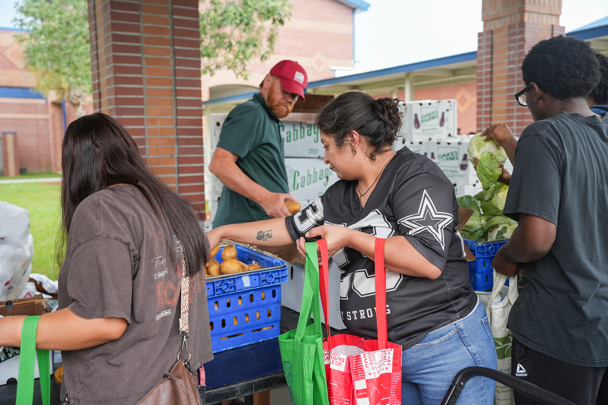 People in outdoor market, picking fresh produce. Three women and a man with bags.