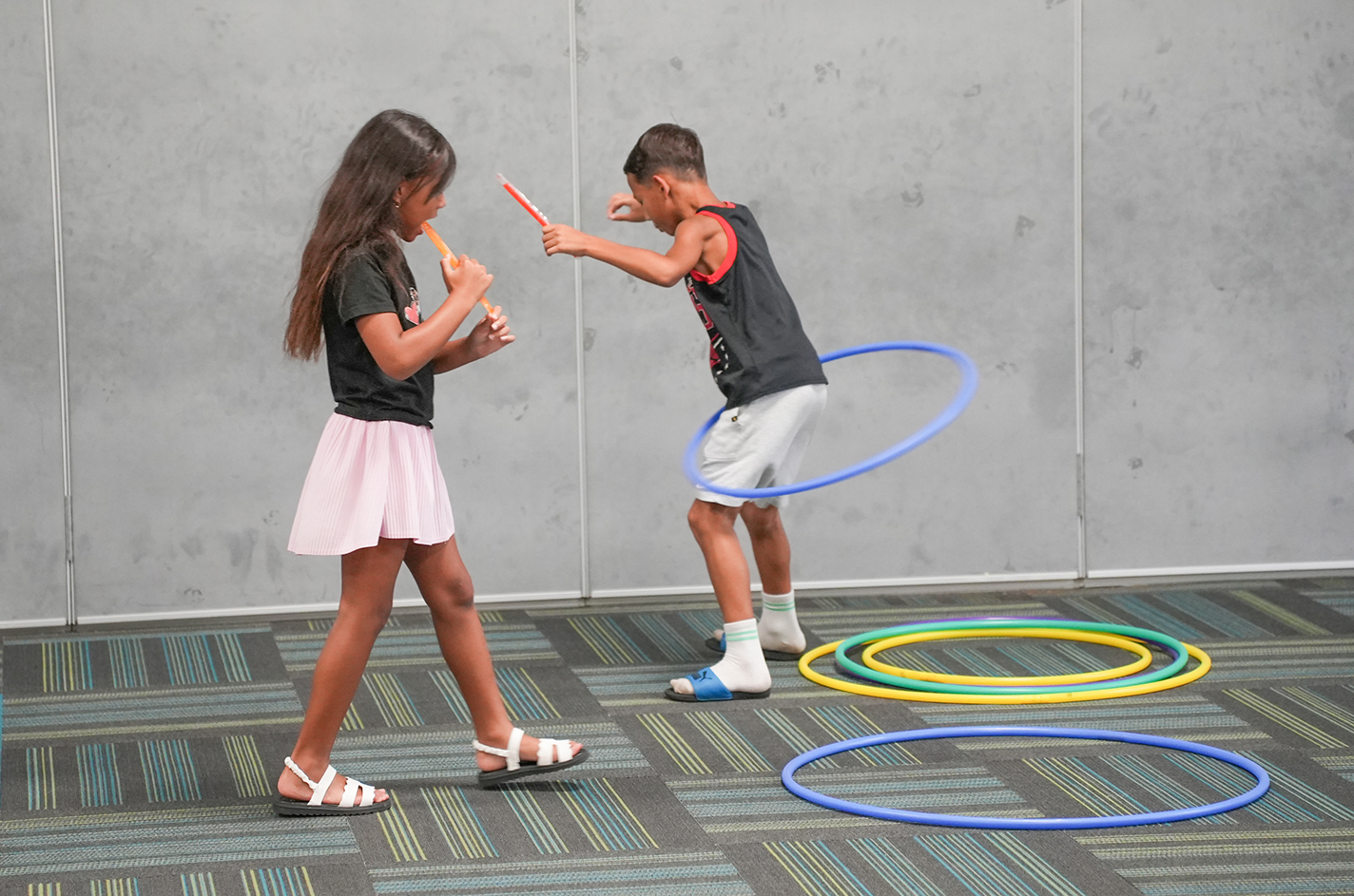 Two children play with hula hoops in an indoor space with a gray wall and patterned floor.