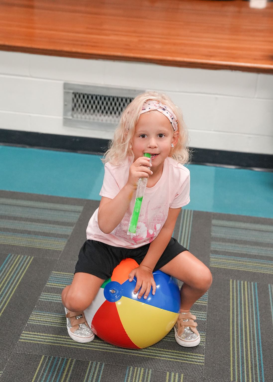 A young girl sits on a colorful beach ball, holding a green tube, possibly blowing bubbles. She wears a pink shirt and black shorts. Behind her, a wooden bench and air vent are visible.