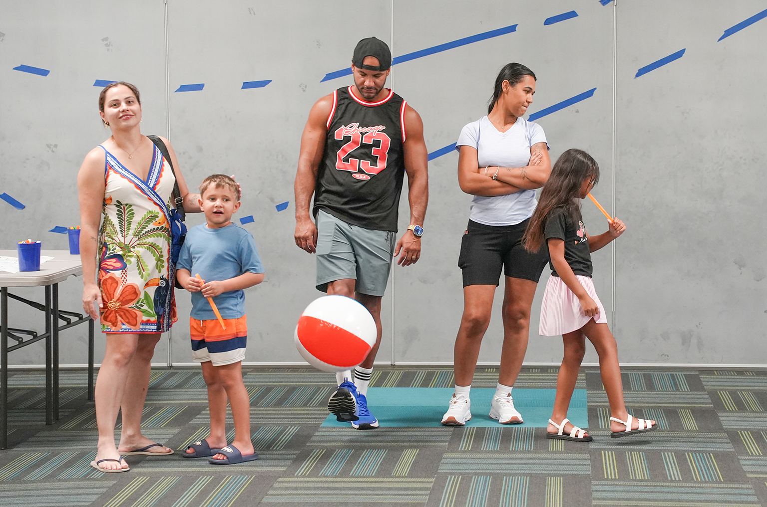 A group of people standing in a room with a ball and a mat. A person in a jersey holds a ball.
