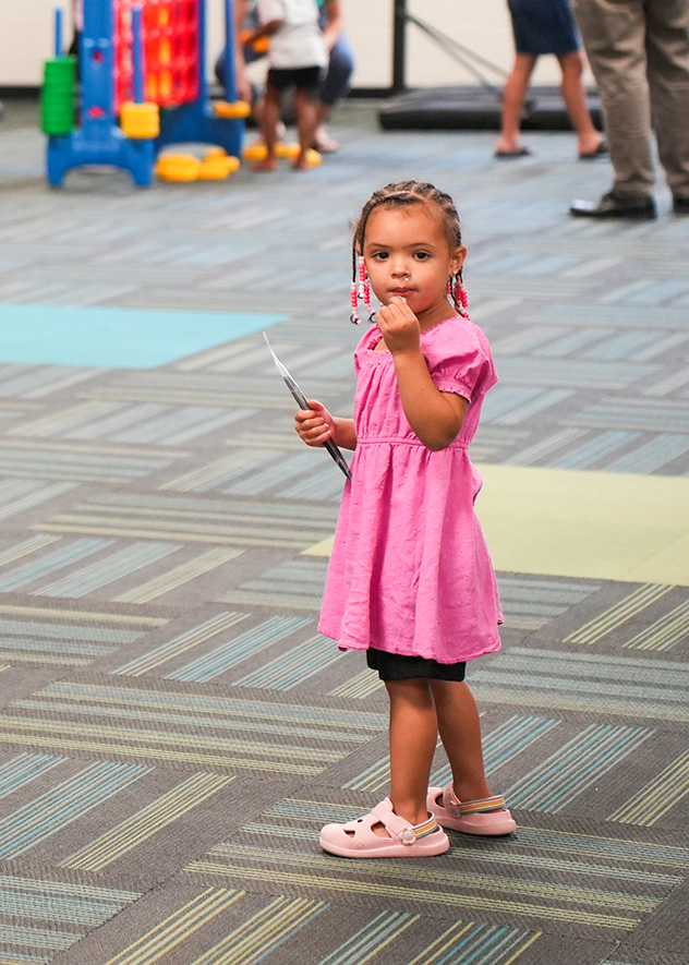 A child stands in a playroom, holding a toy stethoscope, with toys and adults in the background.