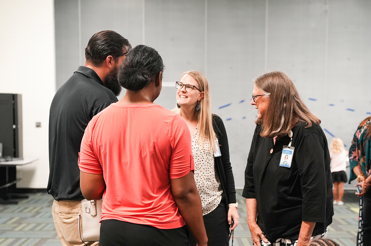 People conversing in an indoor area; one wears glasses, another has a visible ID. Background includes walls, carpet, and a monitor.