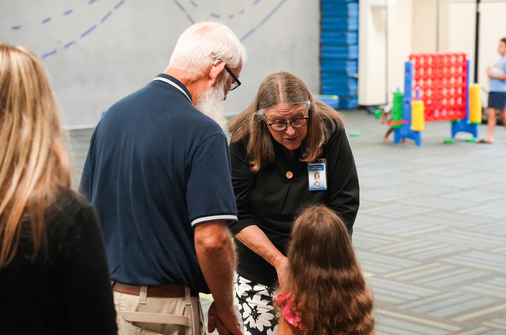 A group of people stands in a room. An older man and woman interact with a young girl.
