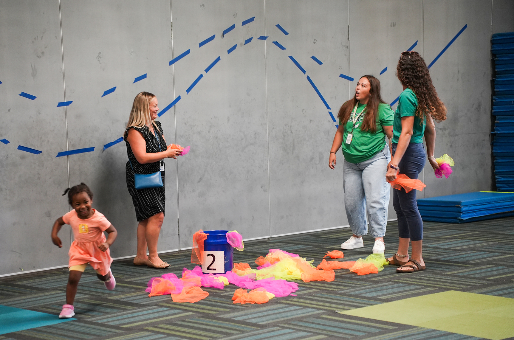 Three women and a child in a gym with multicolored mats. One woman holds a pom-pom.