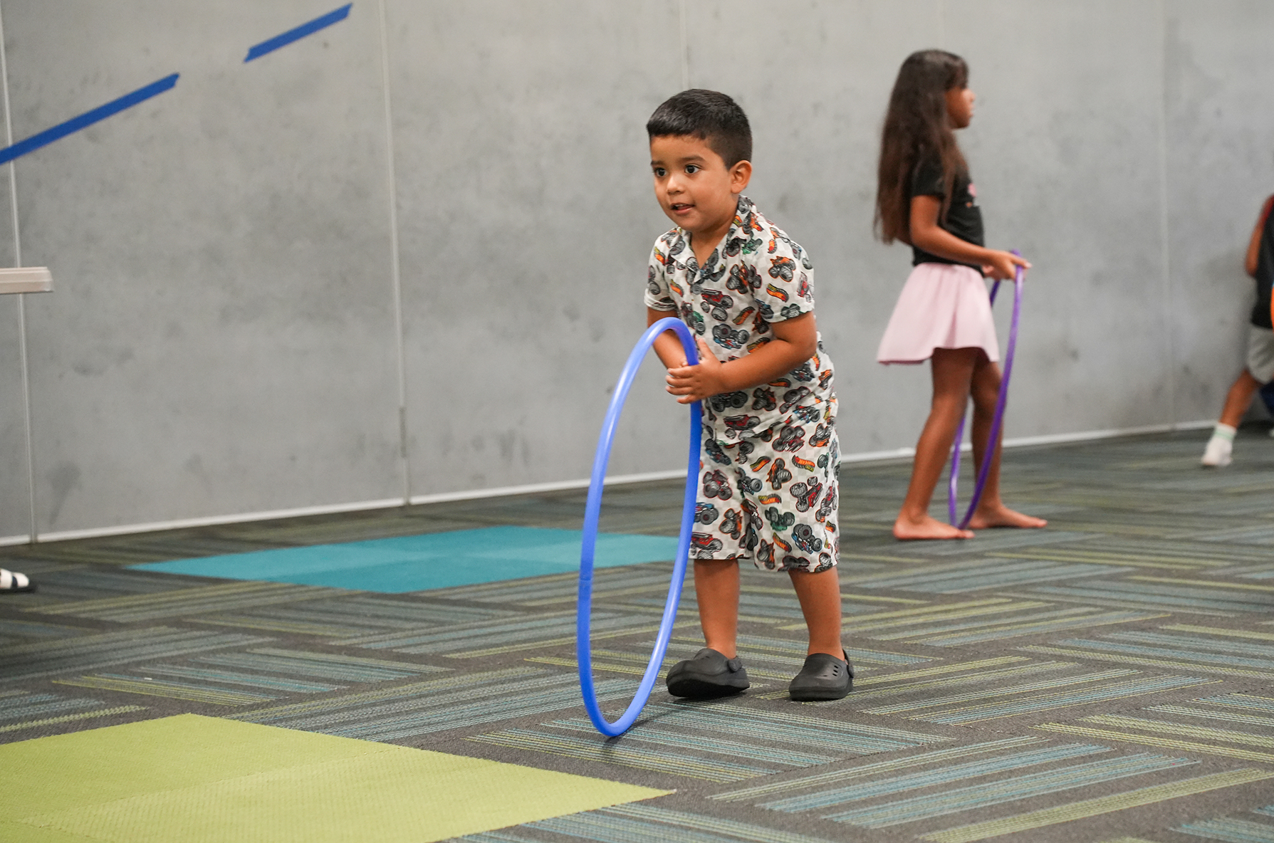 A child in a patterned shirt holds a blue hula hoop in a room with patterned floor. Two girls stand nearby, one holding a purple hula hoop.