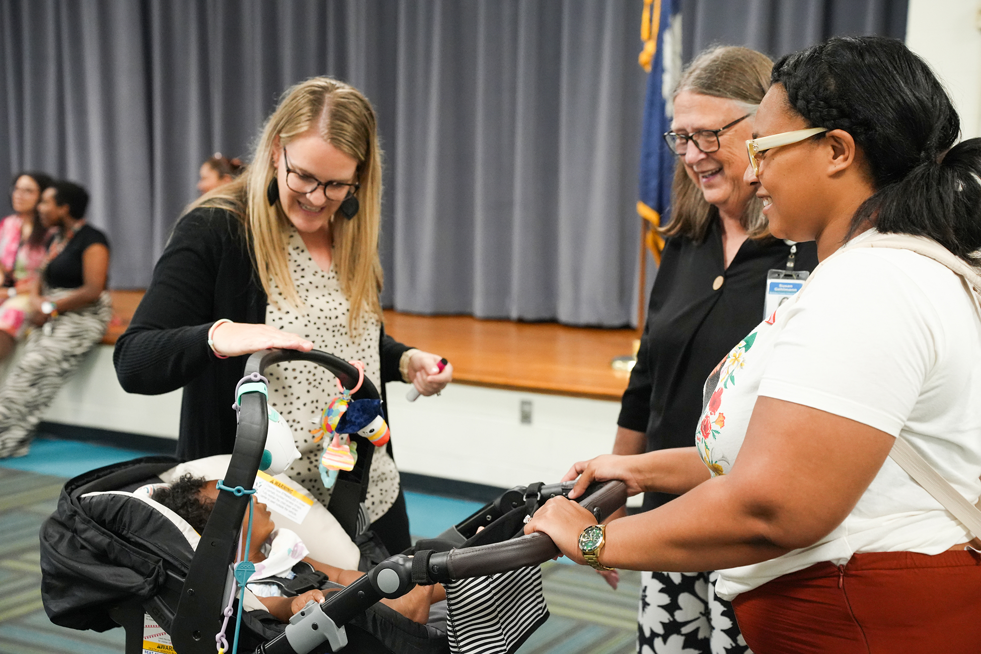 Three women are interacting with a baby in a stroller. A woman in glasses is holding the stroller.