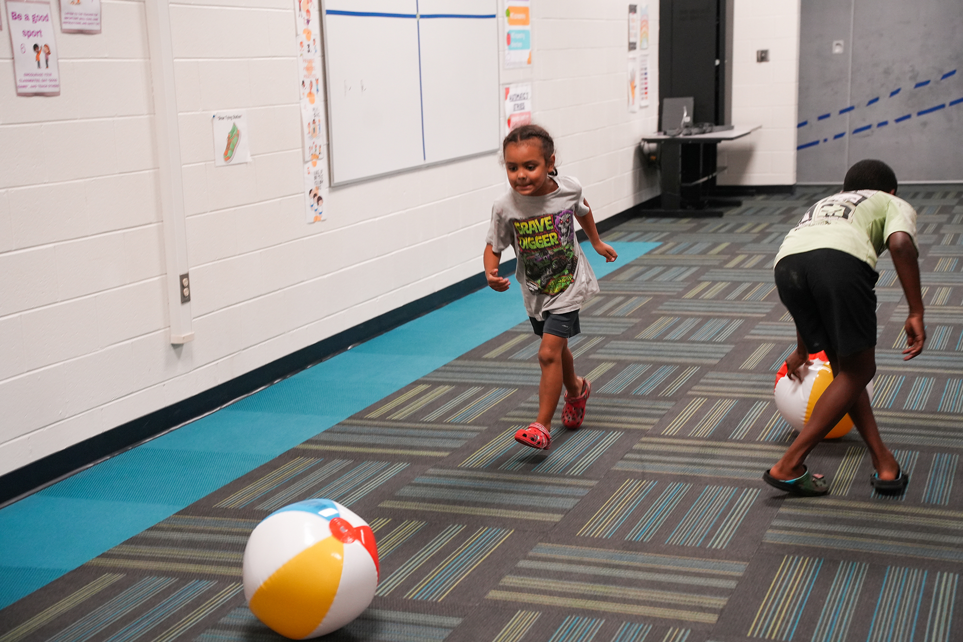 Two children play with large beach balls in a room with white walls, blue flooring, and a whiteboard.