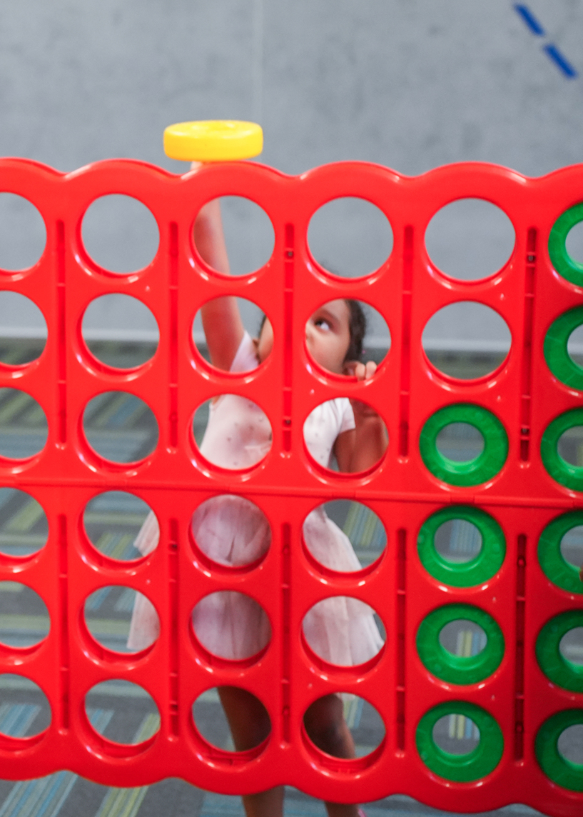 A young child in a pink dress plays with a yellow game chip in a red and green Connect Four game board.