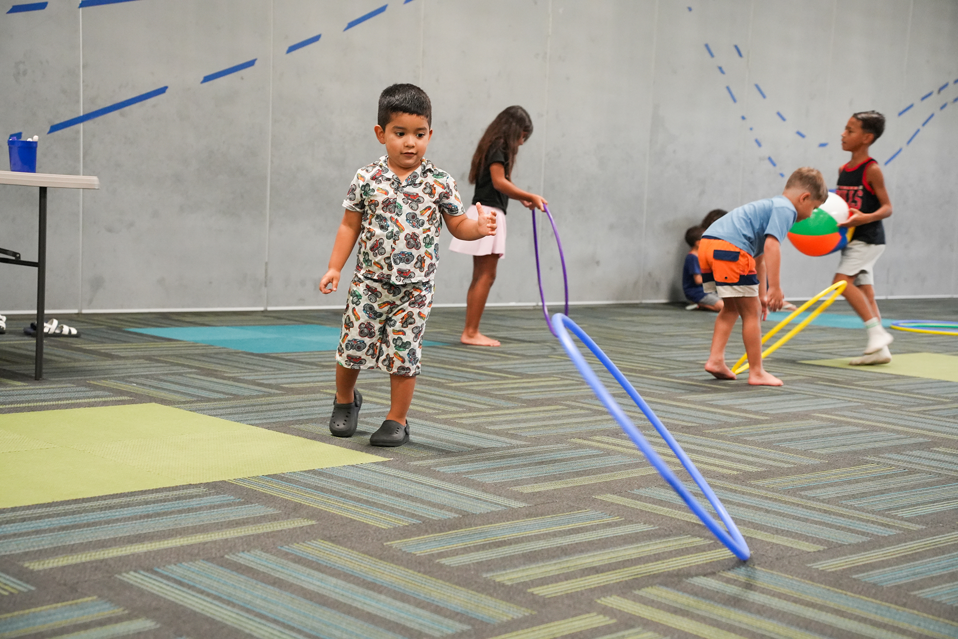 Children play in a gym, with one holding a hula hoop, while others play with balls and a girl holds a hula hoop.
