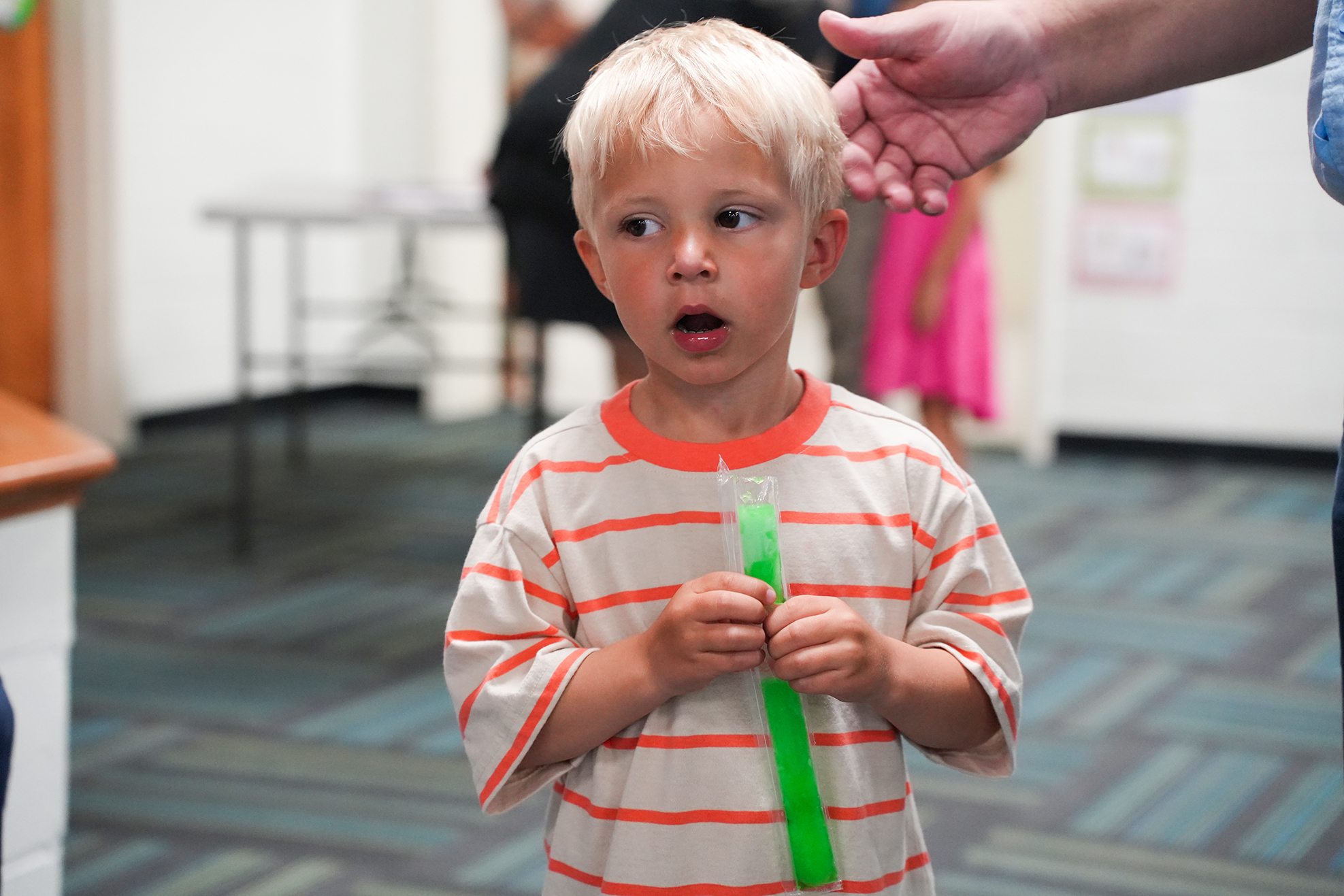 A young boy with blond hair holds a green object while looking upward. A person holds his hand.