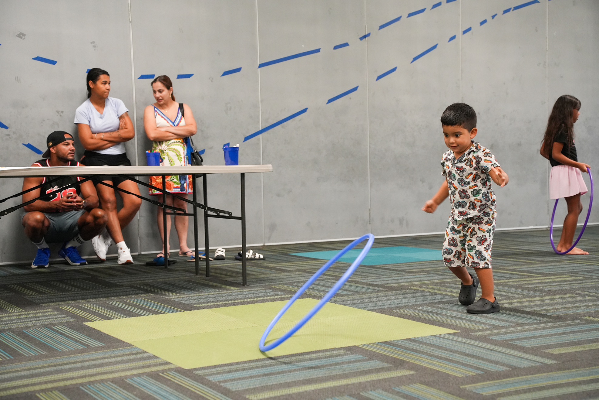 A child wearing shorts and a patterned shirt plays with a blue hula hoop in a room with others watching.